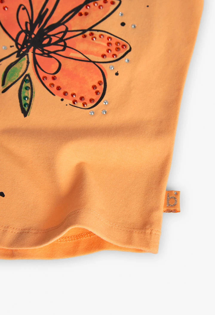 Detalle inferior de blusa naranja con flor ilustrada y aplicaciones brillantes de Boboli.