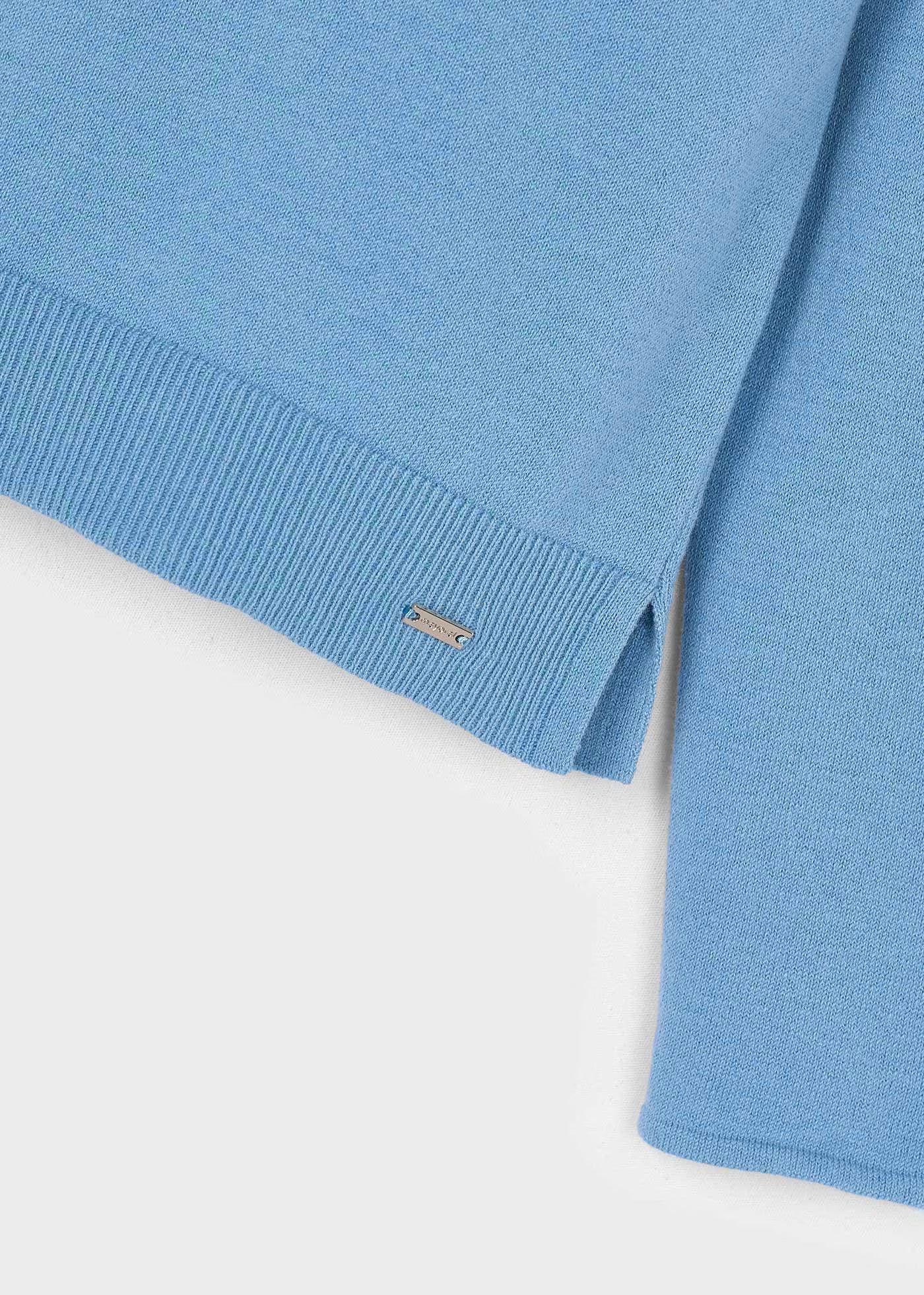 Detalle del bajo y la abertura lateral de un suéter de punto fino color azul, con una placa metálica de la marca Mayoral.