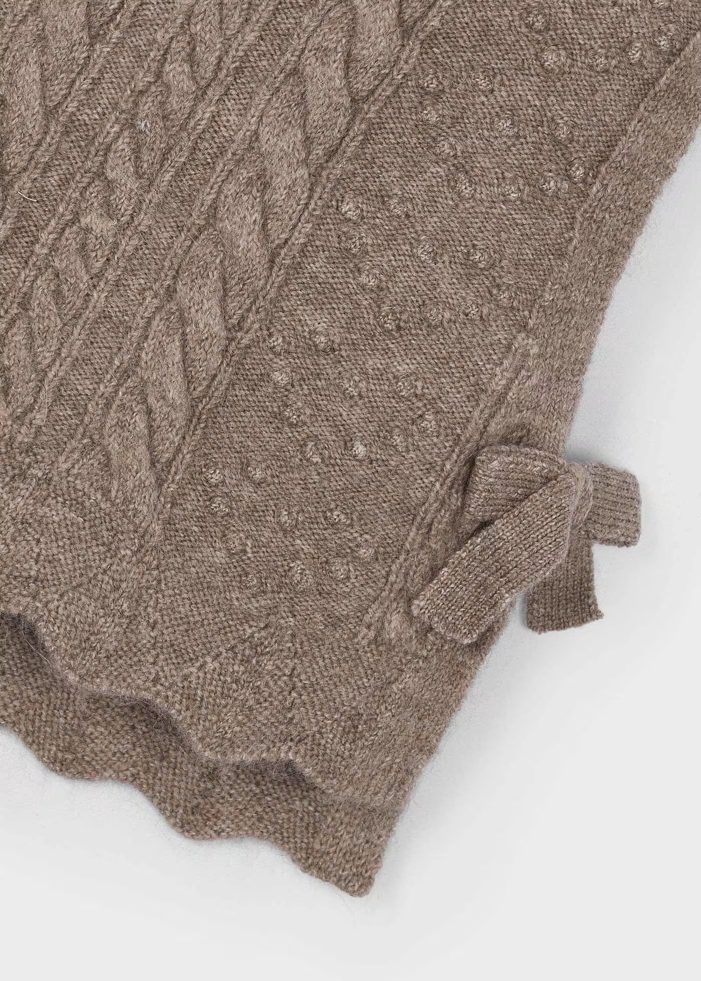 Detalle del tejido trenzado, el bajo ondulado y el moño lateral de un chaleco de punto, marca Mayoral.