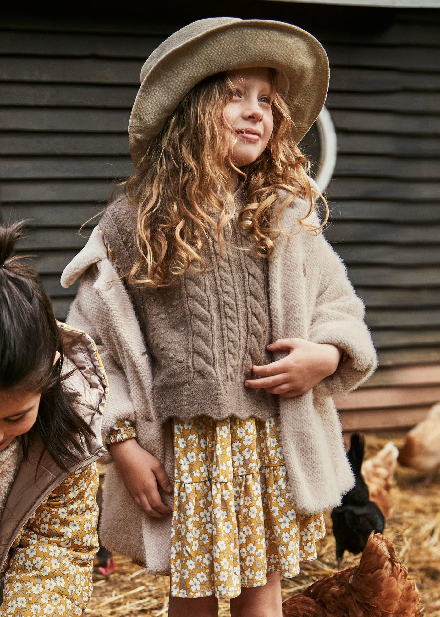 Niña en una granja usando un chaleco de punto trenzado color topo sobre un vestido floreado, marca Mayoral.
