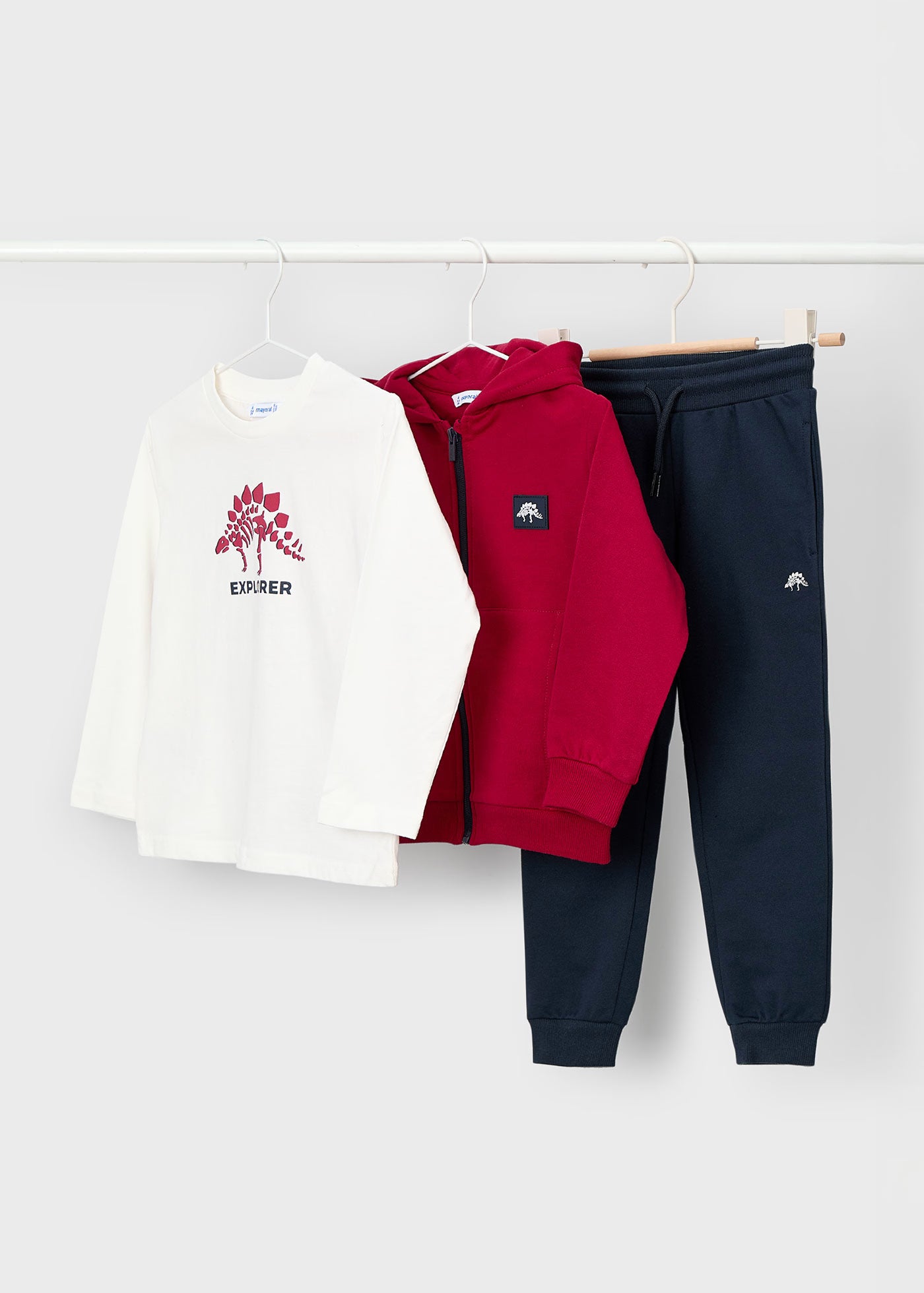Tres piezas de ropa de niño colgadas: una playera blanca con estampado de dinosaurio, una sudadera roja con cremallera y un pantalón jogger azul marino, marca Mayoral.