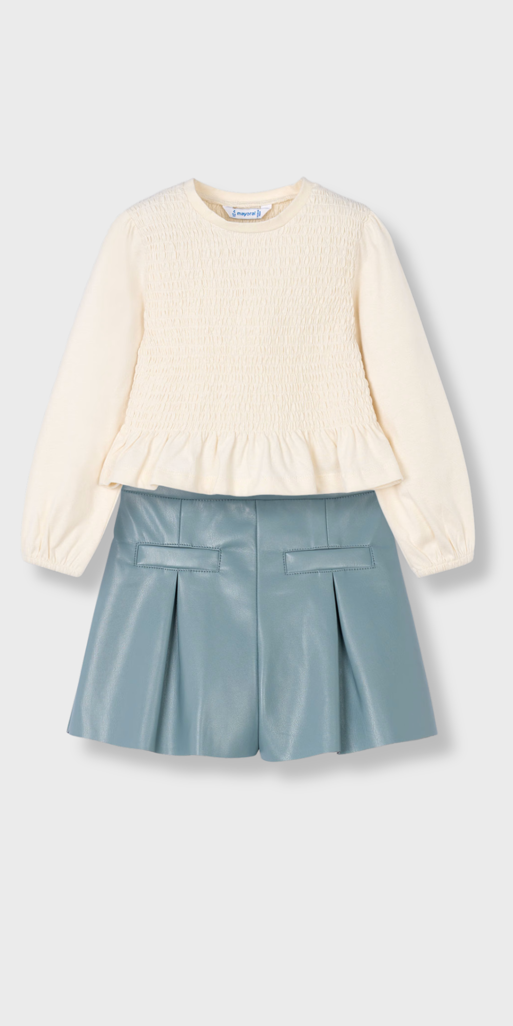 Vista de un conjunto para niña compuesto por una blusa color crudo de nido de abeja y un short de polipiel azul, marca Mayoral.