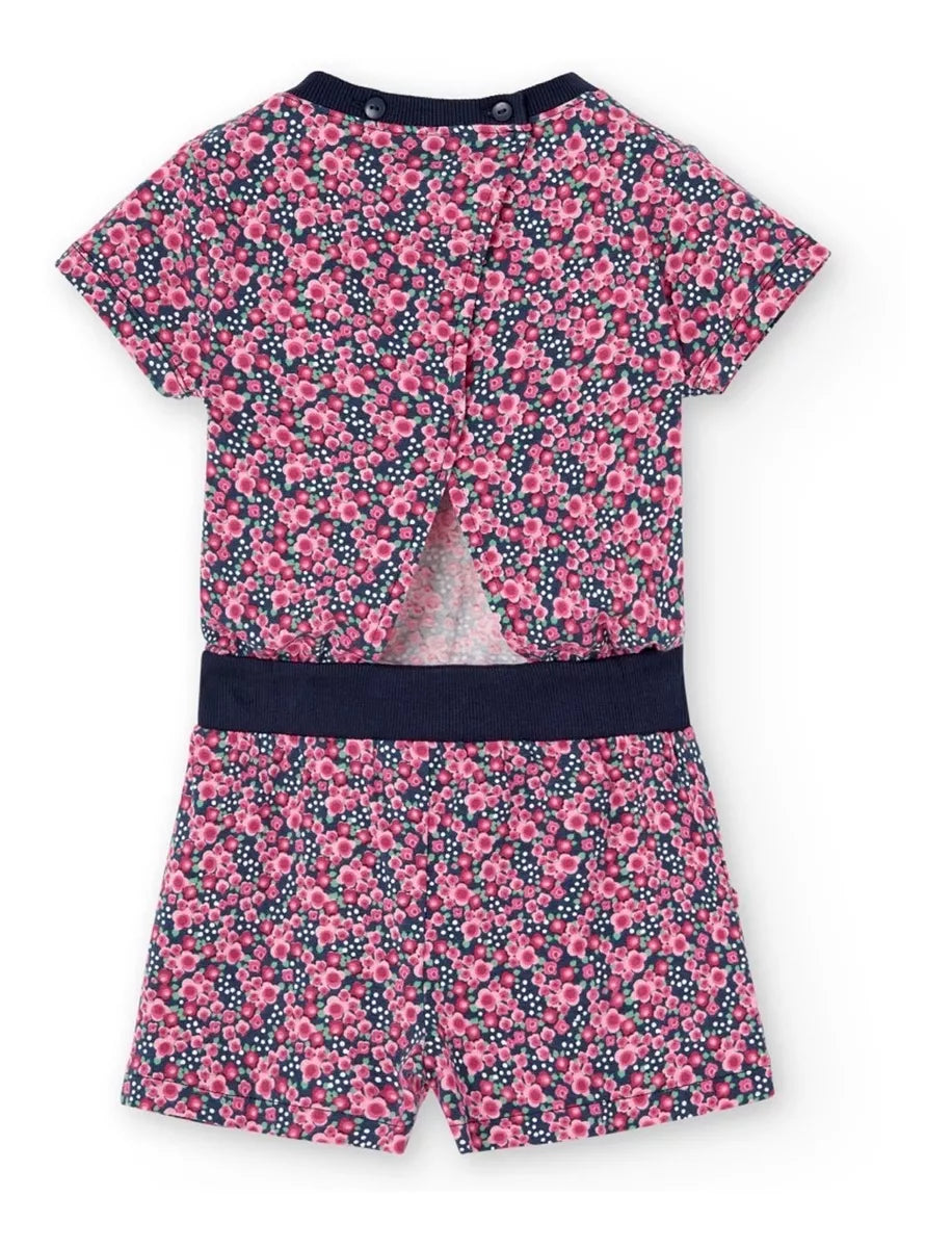 Vista trasera del jumpsuit corto de Boboli con estampado floral en tonos rosas sobre fondo azul marino. Diseño de manga corta con abertura en la espalda y cierre con botones en el cuello. Cintura elástica con banda azul y bolsillos laterales.