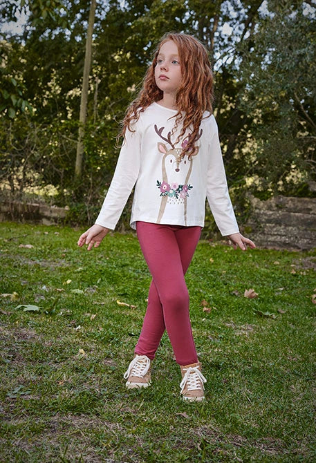 Niña posando en un jardín con jeggings color guinda y una playera blanca con estampado de reno, marca Boboli.