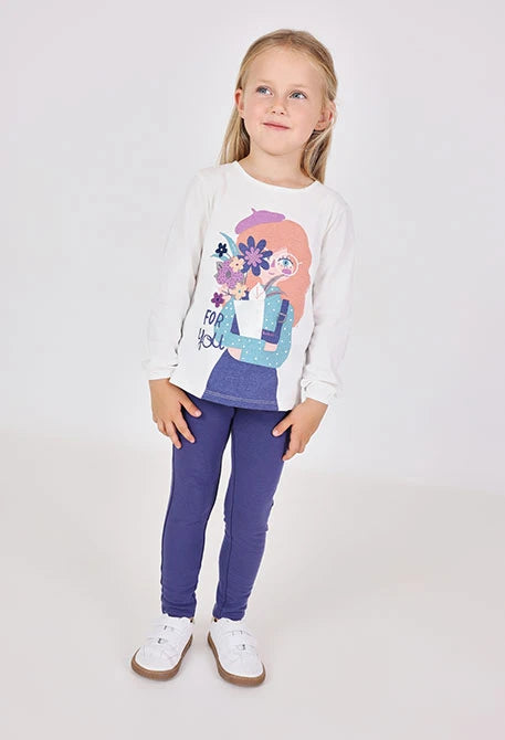 Niña de pie con jeggings color morado y una playera blanca con estampado, marca Boboli.