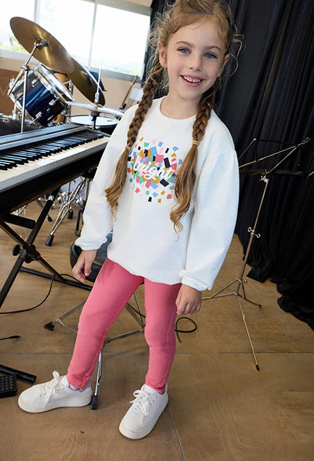 Niña sonriendo en un estudio musical, usando leggings color coral y una sudadera blanca estampada, marca Boboli.