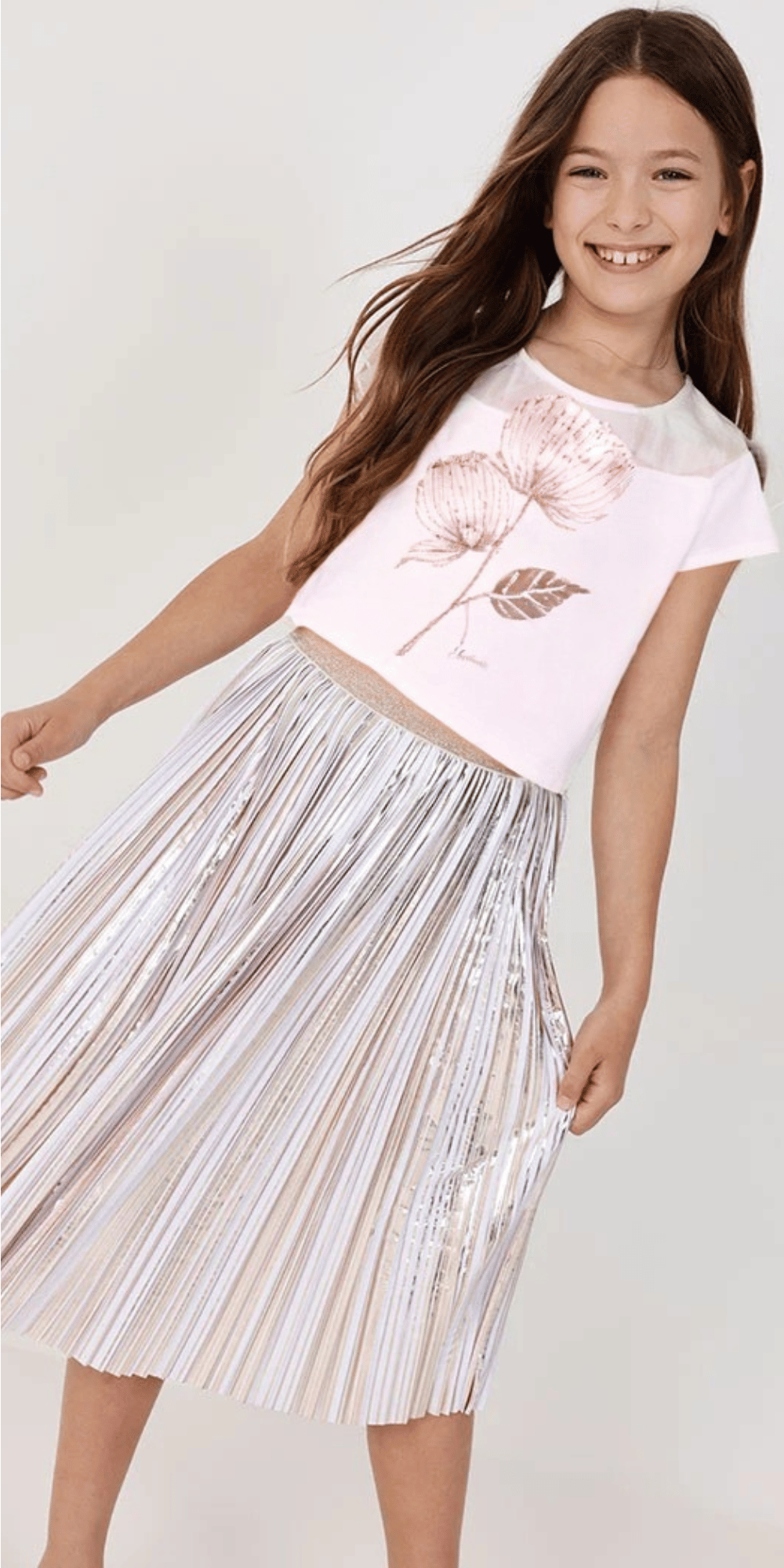 Niña sonriendo con outfit Boboli: playera blanca con flor dorada y falda plisada plateada.