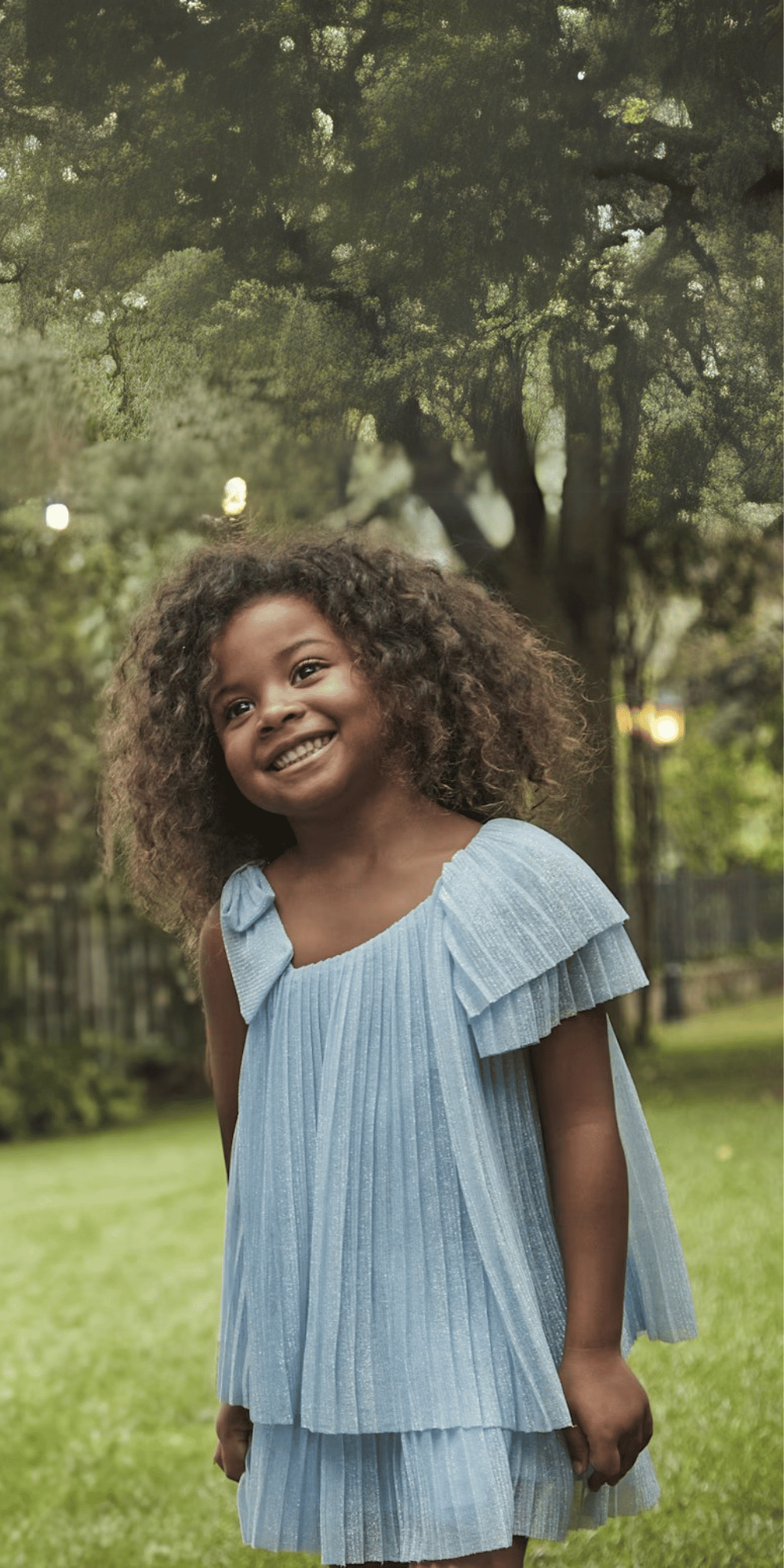 Niña sonriente usando vestido asimétrico plisado en color azul con detalle de una manga con volantes, en un entorno natural.