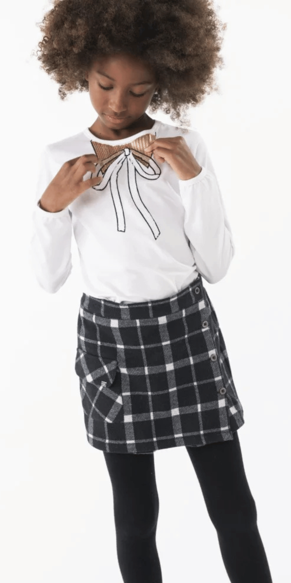 Niña usando conjunto Boboli con falda de cuadros y playera blanca con moño de lentejuelas doradas