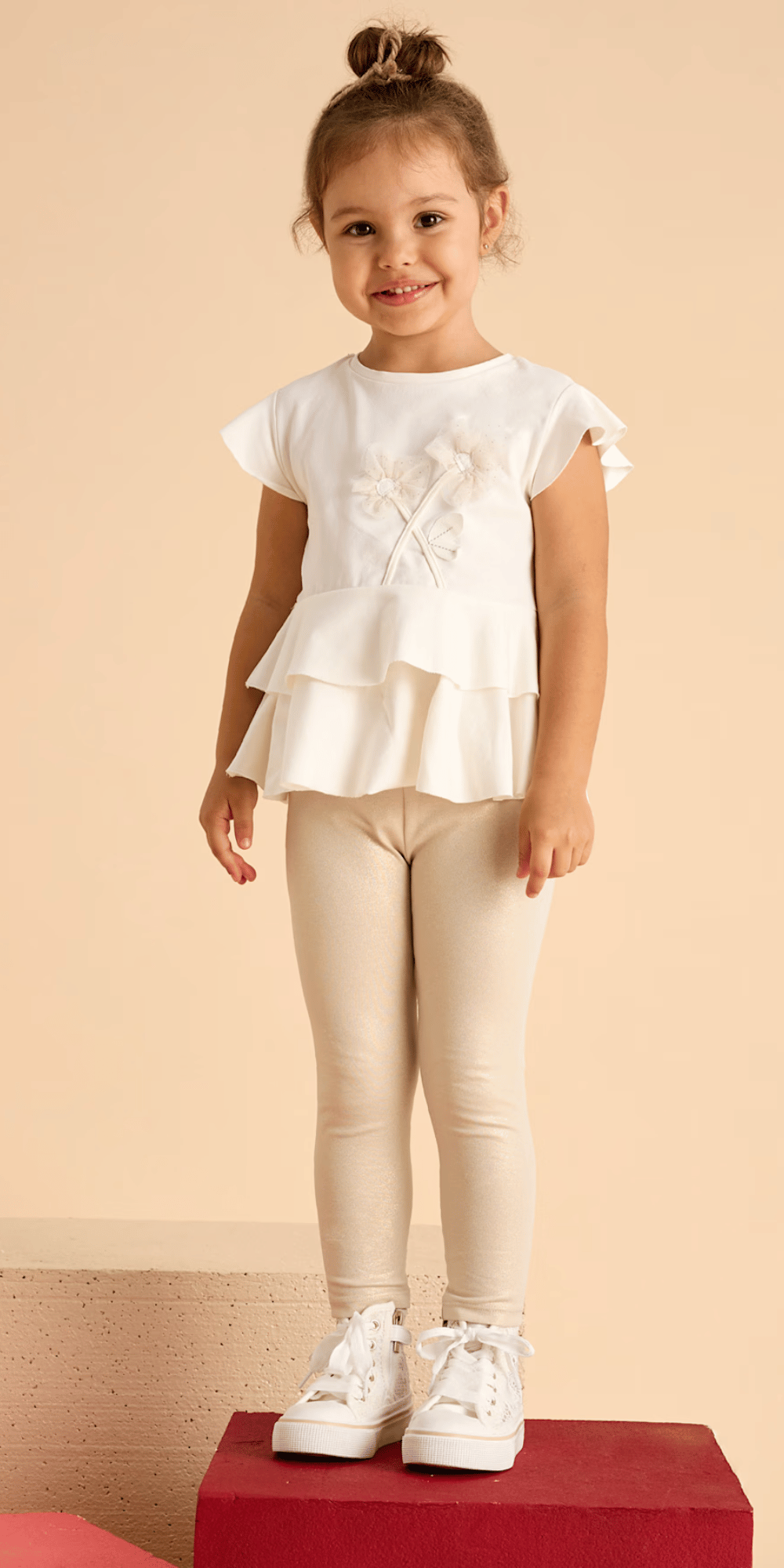 Niña sonriente usando un conjunto de playera con flores y leggings brillosos en tono beige de la marca Mayoral.