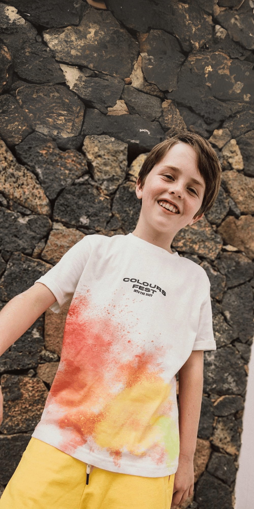 Niño sonriendo con playera blanca de estampado de festival de colores y pantalones cortos amarillos.