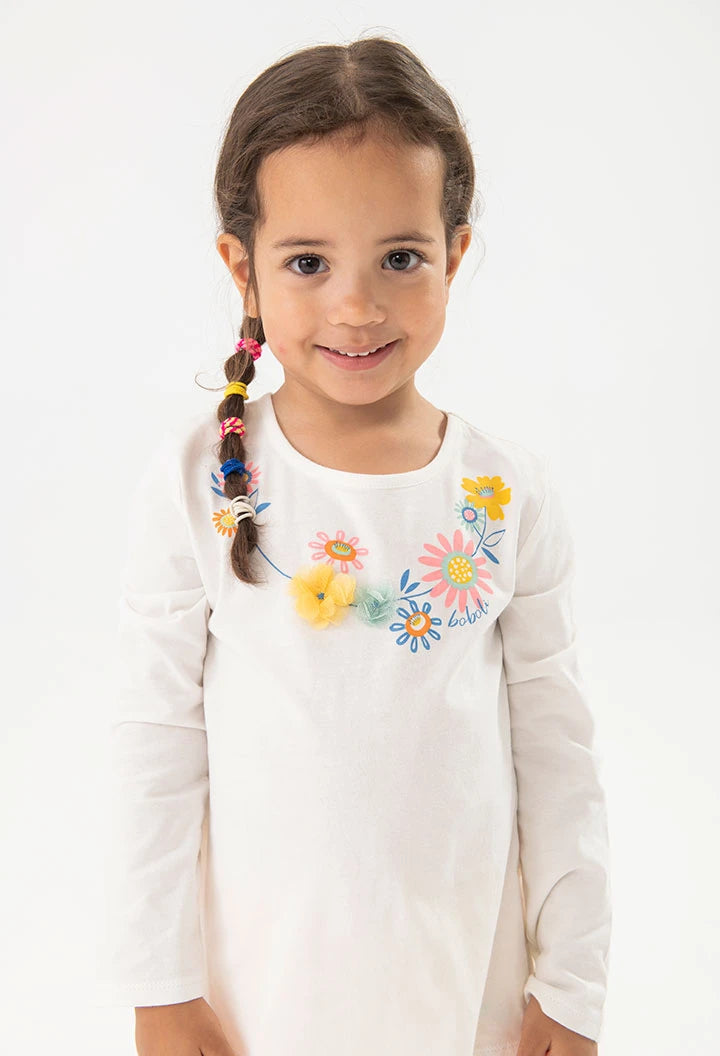 Niña sonriente con cabello trenzado, usando una playera blanca de manga larga con detalles florales bordados de colores.