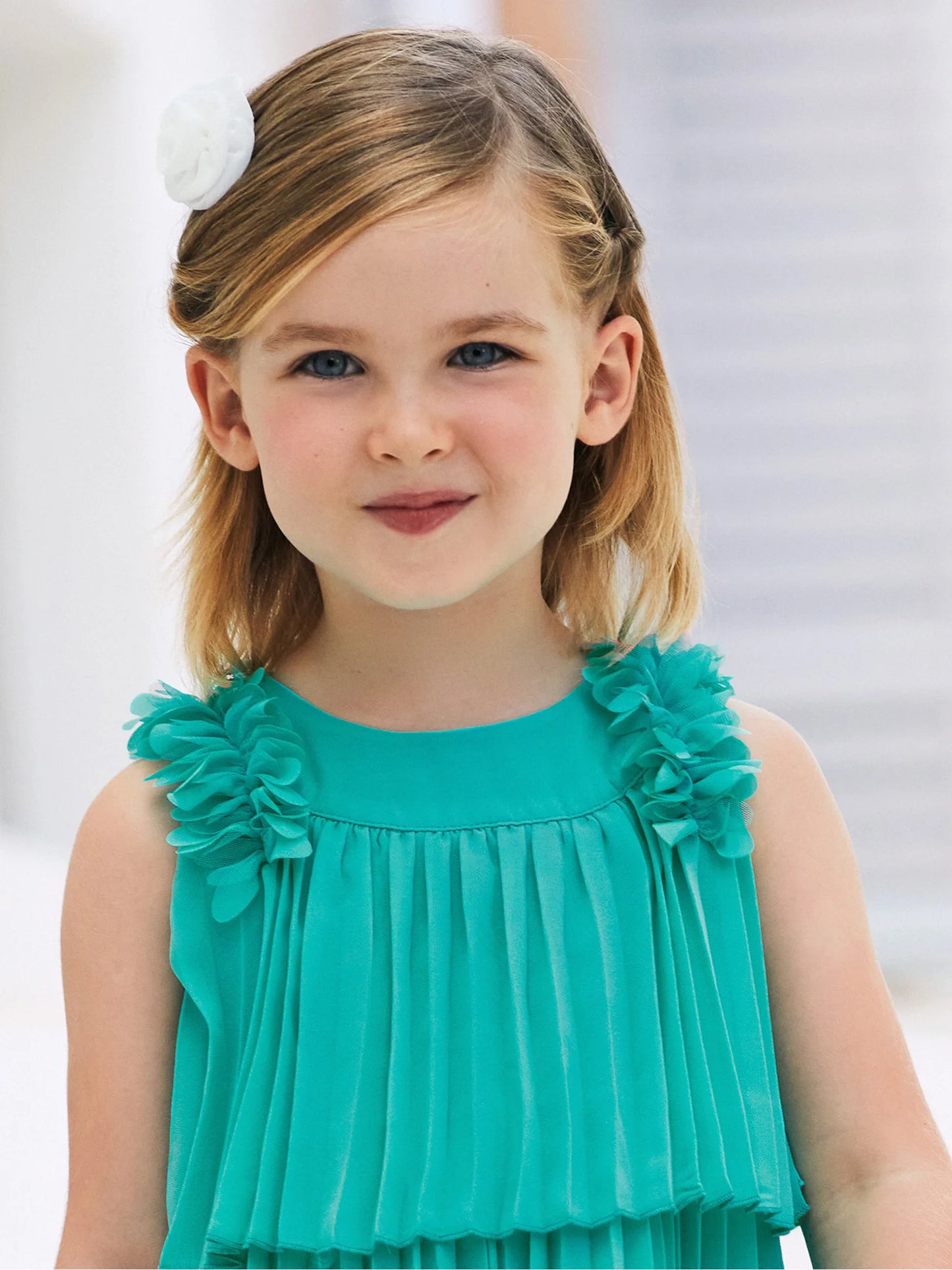 Primer plano de niña usando un vestido plisado en color verde con detalles de flores en los hombros. Diseño sofisticado ideal para eventos especiales y ceremonias.