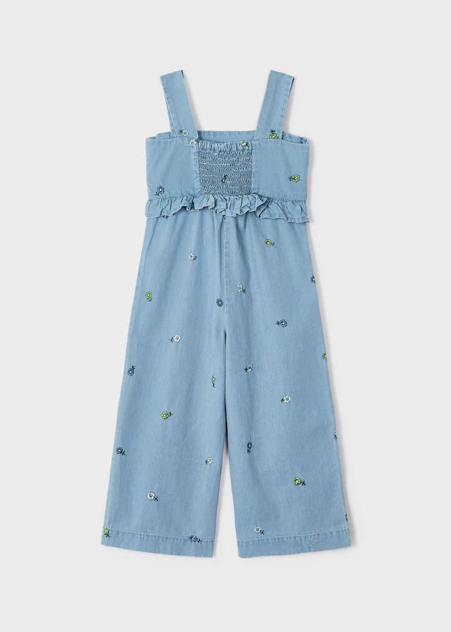 Jumpsuit de mezclilla azul claro para niña con bordado floral en todo el conjunto, espalda con detalle de smock elástico, volantes y tirantes anchos. Diseño fresco y versátil ideal para cualquier ocasión.