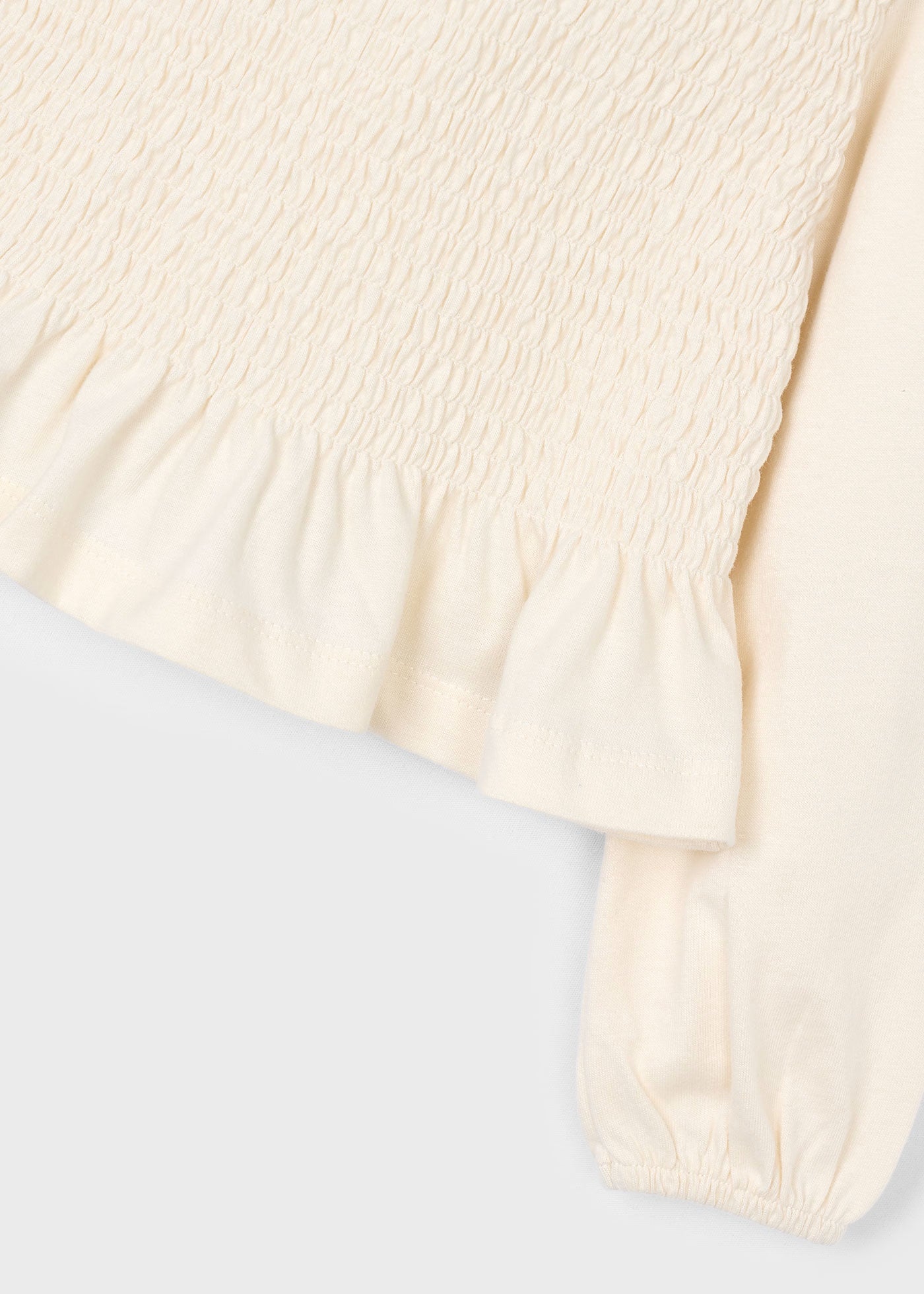 Detalle del tejido elástico de nido de abeja y el volante en el bajo de una blusa color crudo.