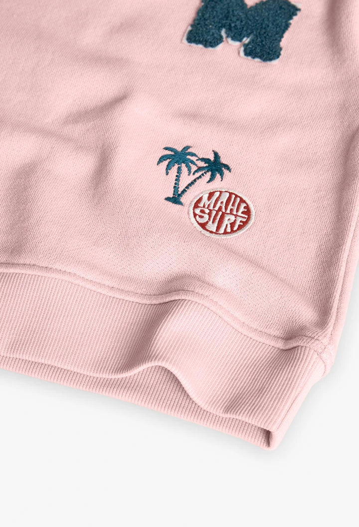Detalle de parches bordados de palmeras y texto “Mahe Surf” en sudadera rosa.