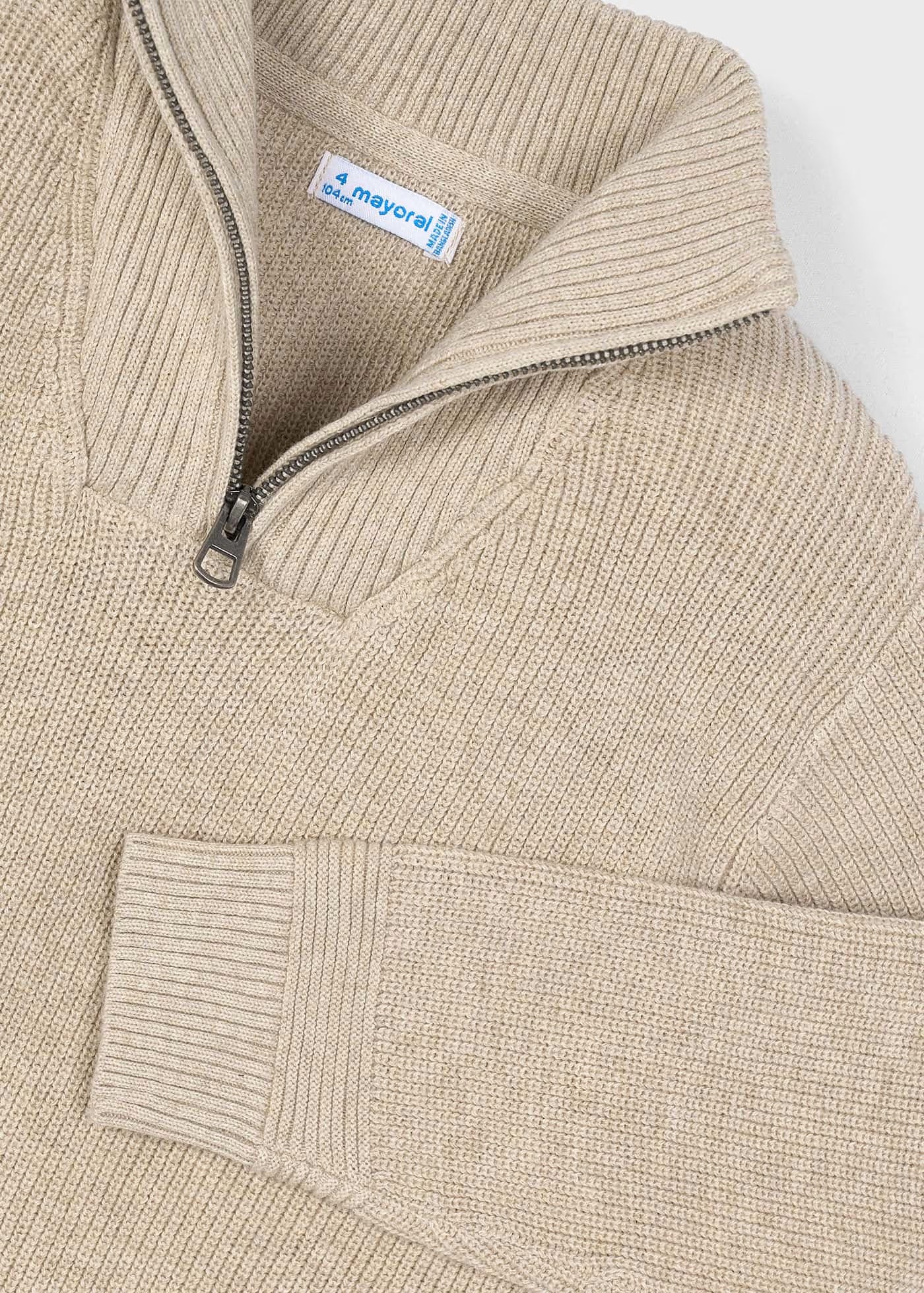 Detalle del cuello y la cremallera de un suéter de punto canalé color beige, mostrando la etiqueta de la marca Mayoral.