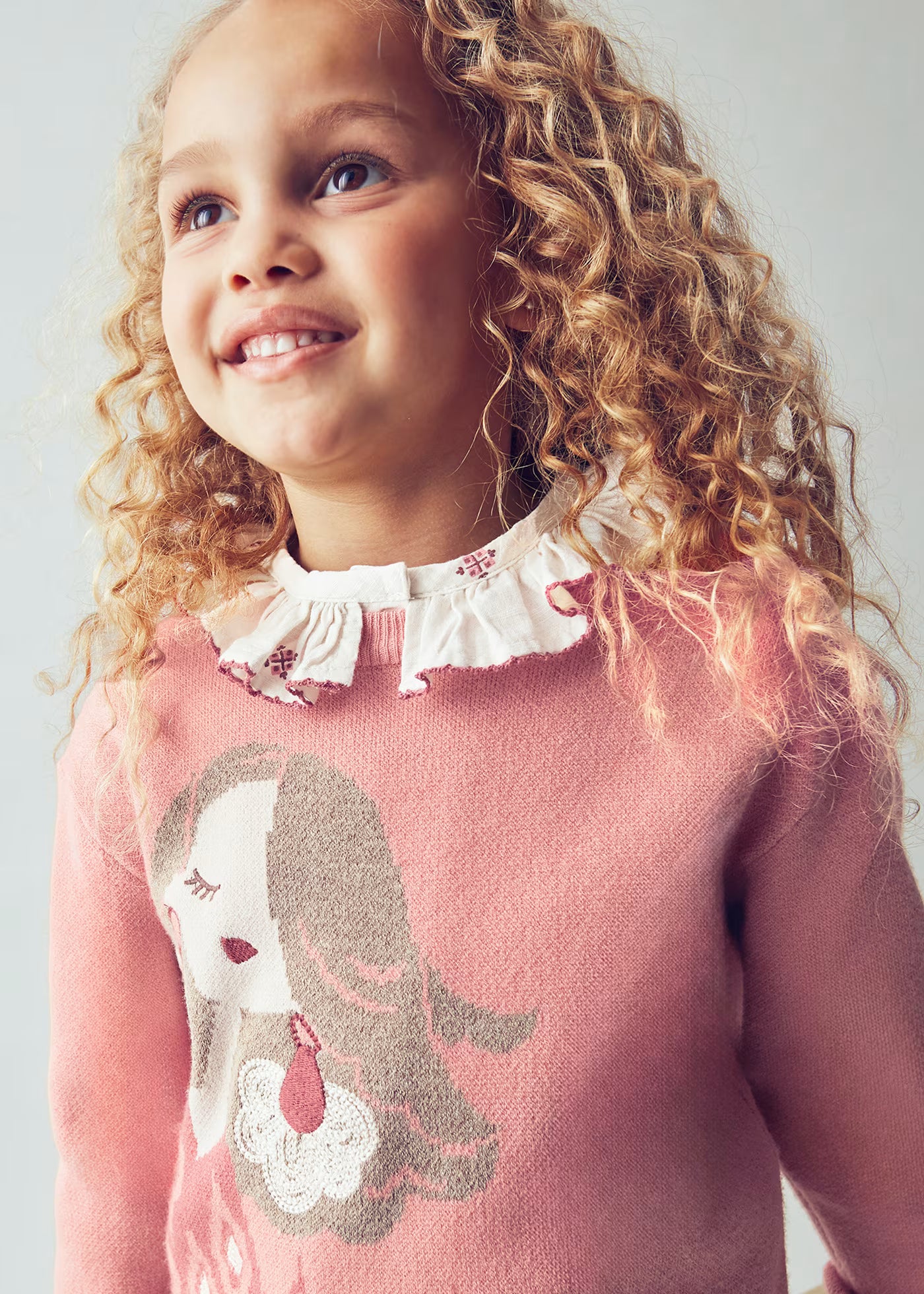 Primer plano de una niña sonriendo, luciendo un suéter rosa con el dibujo de una muñeca y una blusa de cuello con volantes debajo.