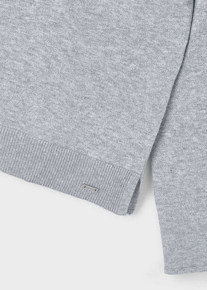 Detalle del bajo y la abertura lateral de un suéter de punto fino color gris, con una placa metálica de la marca Mayoral. 