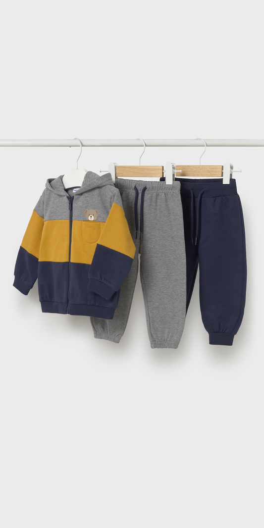 Tres piezas de ropa de bebé colgadas: una sudadera con capucha de osito y dos pantalones jogger, uno gris y uno azul marino, marca Mayoral.