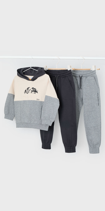 Tres piezas de ropa de niño colgadas: una sudadera con capucha y diseño de montaña, y dos pantalones jogger en gris oscuro y gris claro, marca Mayoral.