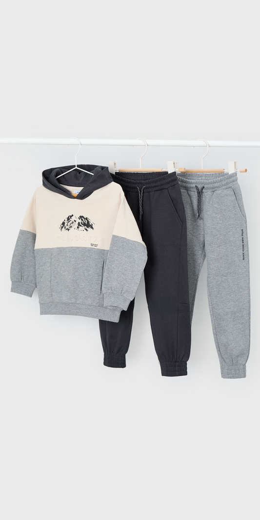 Tres piezas de ropa de niño colgadas: una sudadera con capucha y diseño de montaña, y dos pantalones jogger en gris oscuro y gris claro, marca Mayoral.