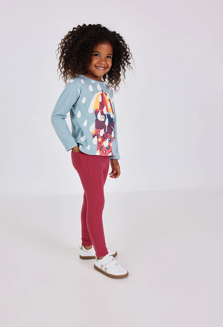 Niña de pie sonriendo con jeggings color guinda y una playera azul claro con estampado de paraguas, marca Boboli.
