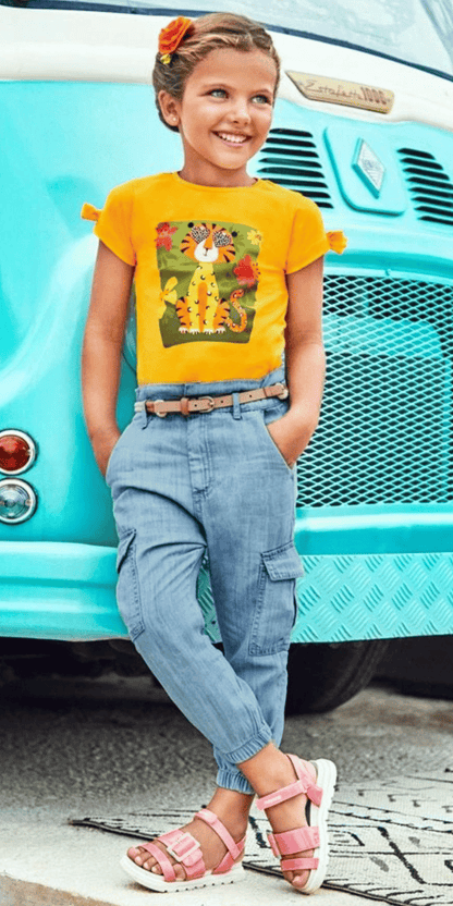 Niña con outfit casual compuesto por blusa amarilla con estampado de tigre, pantalón cargo denim y sandalias rosas, posando junto a una camioneta vintage en un look moderno y relajado.