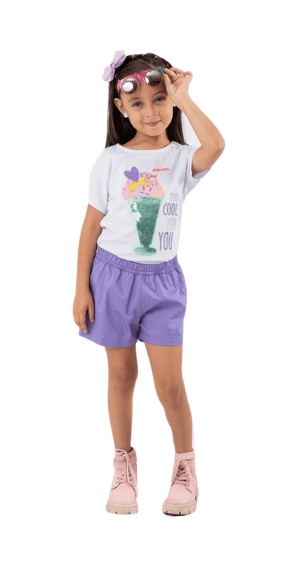 Niña con outfit infantil compuesto por una blusa blanca con estampado de milkshake y lentejuelas reversibles con la frase “Too Cool For You”, shorts morados, botines rosas y gafas de sol en la cabeza, creando un look veraniego y moderno.