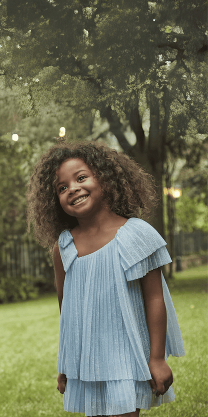 Niña sonriente usando vestido asimétrico plisado en color azul con detalle de una manga con volantes, en un entorno natural.