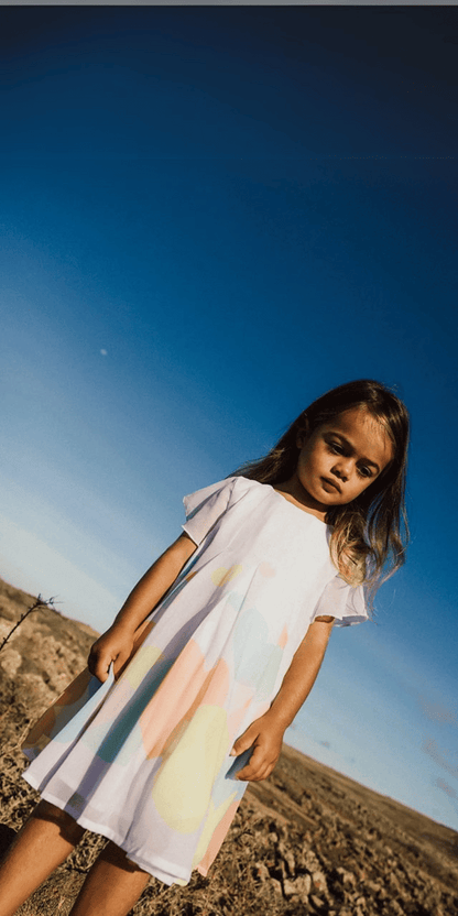 Niña vistiendo un vestido blanco con estampado de corazones de colores pastel en un paisaje natural con cielo azul de fondo.