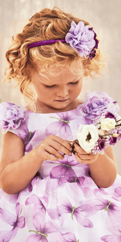 Niña con vestido de fiesta en tonos lila con estampado de mariposas y detalles florales en los hombros, sosteniendo un pequeño ramo de flores. Un look encantador y elegante para ocasiones especiales.