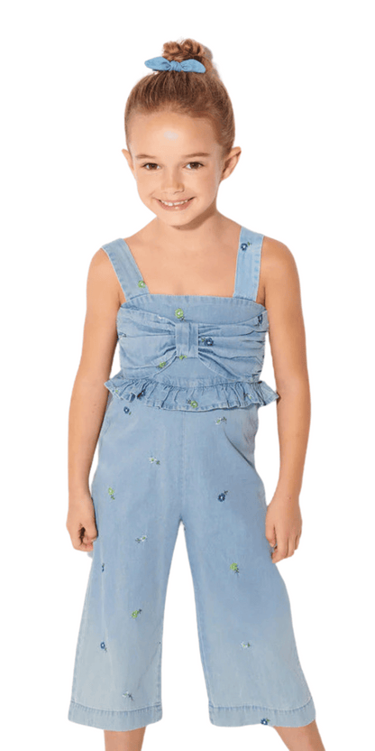Niña sonriendo con un jumpsuit de mezclilla azul claro con tirantes anchos, detalle de lazo y volantes en la parte superior, y bordado floral en todo el conjunto. Outfit cómodo y moderno para niñas.