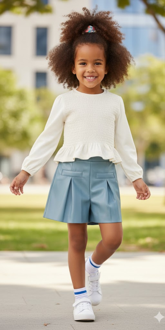 Niña con conjunto compuesto por una blusa color crudo de nido de abeja y un short de polipiel azul, marca Mayoral.