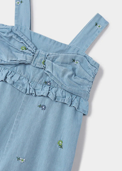 Detalle de jumpsuit de mezclilla azul claro para niña con bordado floral, moño decorativo en el pecho y volantes en el top. Diseño con tirantes anchos y un estilo fresco y cómodo para cualquier ocasión.