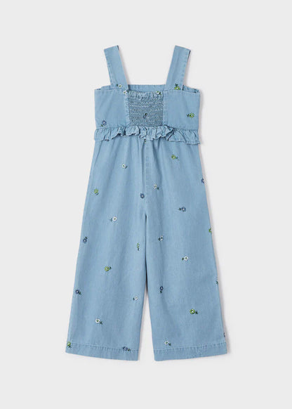 Jumpsuit de mezclilla azul claro para niña con bordado floral en todo el conjunto, espalda con detalle de smock elástico, volantes y tirantes anchos. Diseño fresco y versátil ideal para cualquier ocasión.