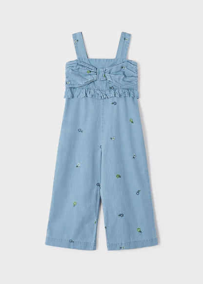 Jumpsuit de mezclilla azul claro para niña con tirantes anchos, detalle de lazo y volantes en el pecho, y bordado floral en todo el conjunto. Diseño moderno y cómodo para cualquier ocasión.