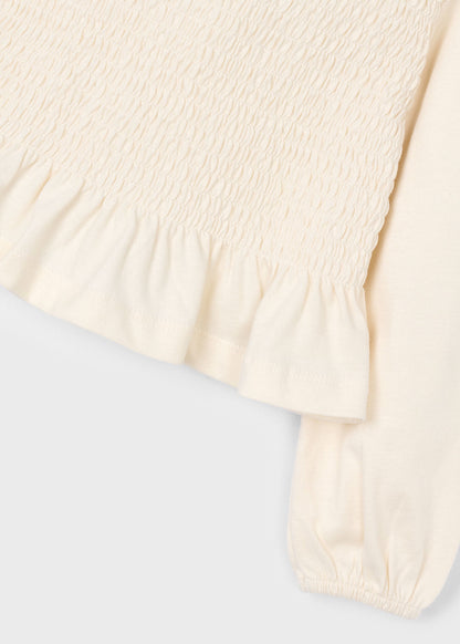 Detalle del tejido elástico de nido de abeja y el volante en el bajo de una blusa color crudo.
