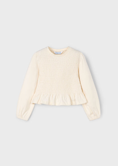 Vista frontal de una blusa de manga larga para niña en color crudo, con cuerpo elástico de nido de abeja y volante.