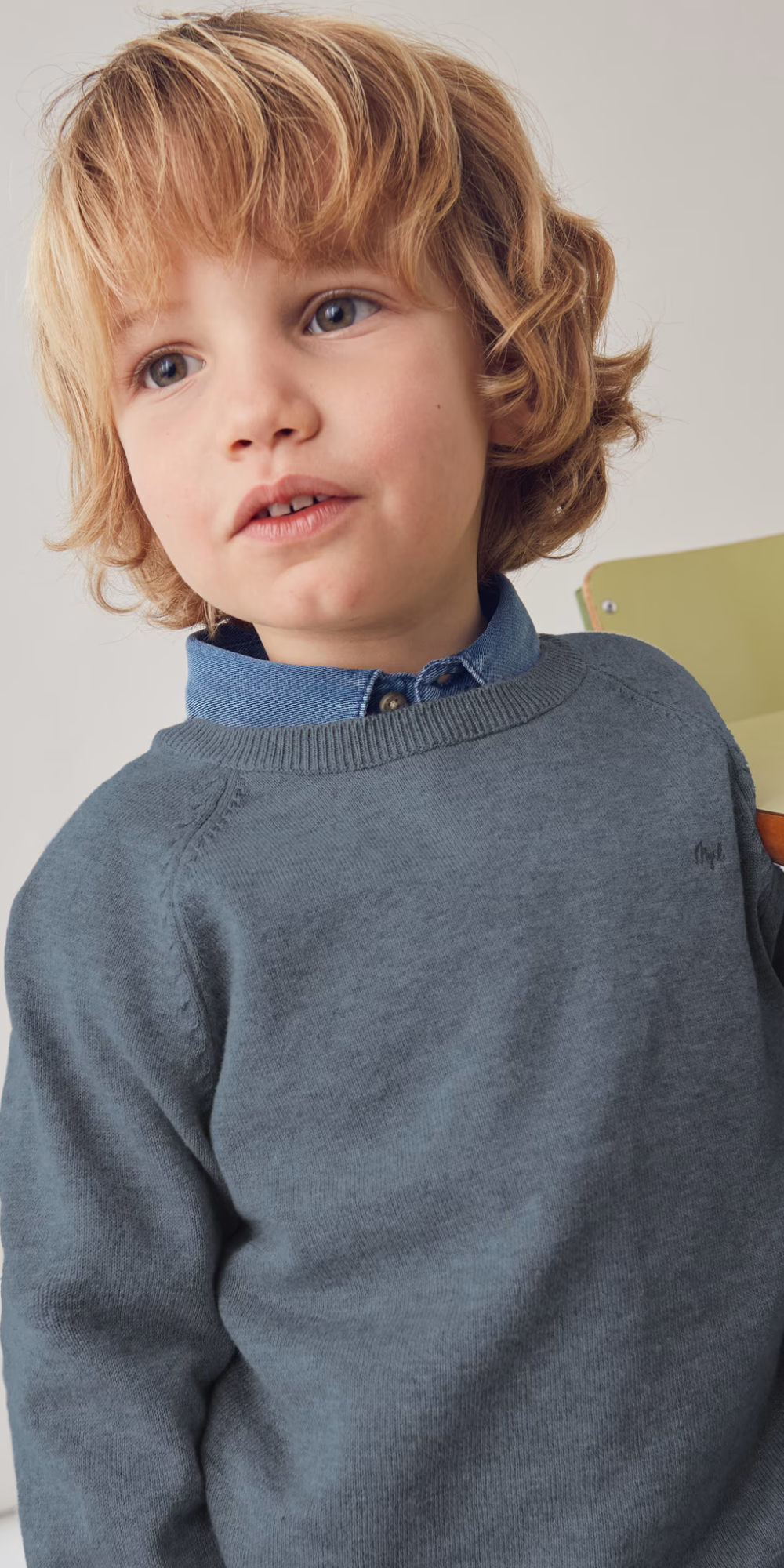 Niño de pelo rubio vistiendo el suéter gris Mayoral sobre una camisa de mezclilla azul.