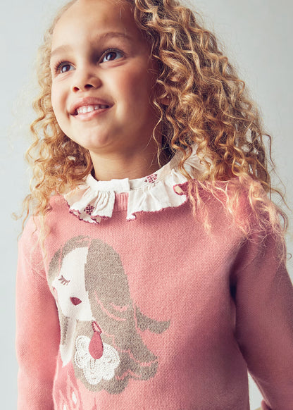 Primer plano de una niña sonriendo, luciendo un suéter rosa con el dibujo de una muñeca y una blusa de cuello con volantes debajo.