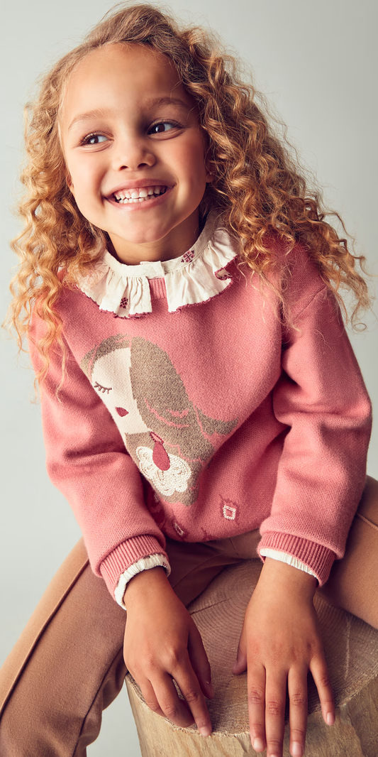Niña sonriendo sentada en un tronco, usando un suéter rosa con el dibujo de una muñeca, combinado con una blusa de cuello con olanes.