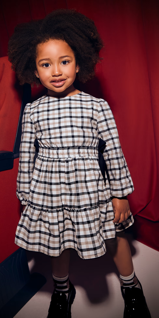 Niña de pie junto a una butaca de cine, usando un vestido de manga larga con estampado de cuadros y moños en la cintura, marca Mayoral.