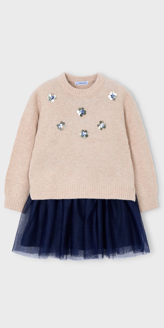Vista frontal de un conjunto de dos piezas para niña: un suéter de punto color avena con flores de lentejuelas sobre un vestido con falda de tul azul marino, marca Mayoral.